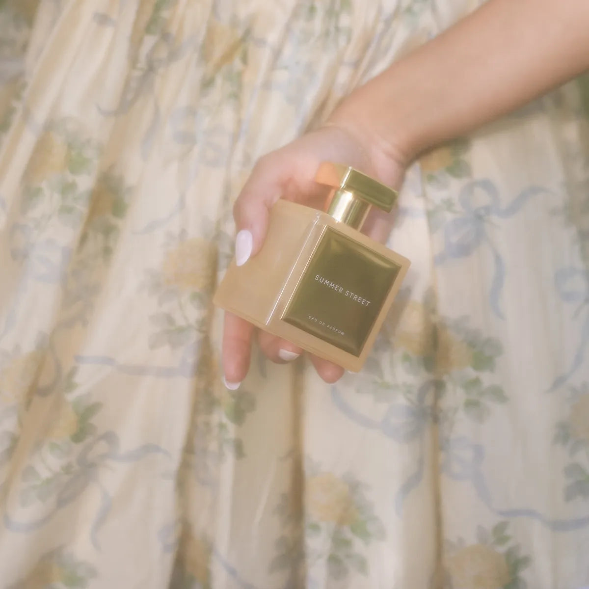Summer Street Eau de Parfum by House of Brandt held in hand against soft floral dress background, elegant amber glass perfume bottle with gold cap lifestyle shot