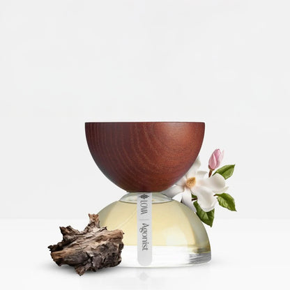 Agonist by LOVA niche perfume bottle with sculpted wooden cap, pale golden fragrance, white magnolia flower, and natural wood accent