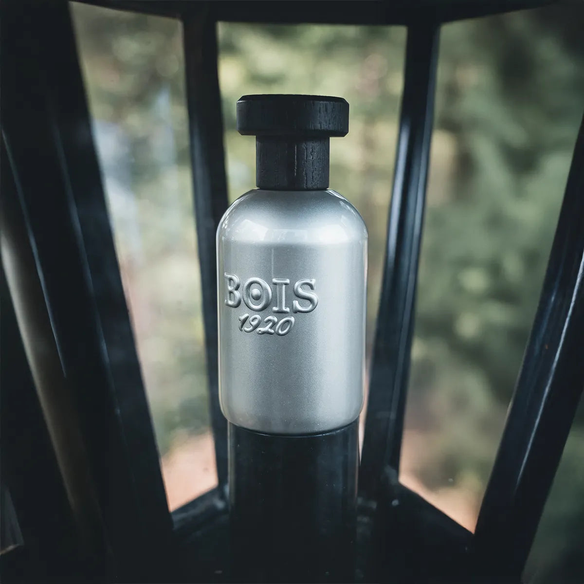 Bois 1920 Dolce Di Giorno perfume bottle with metallic silver finish and black cap placed inside a lantern with soft natural background.