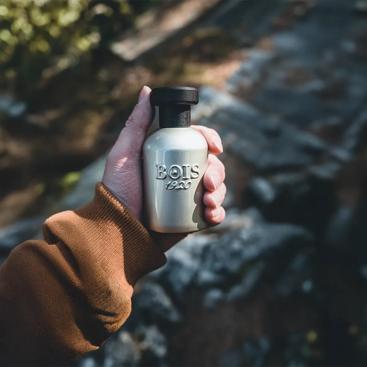 Hand holding Bois 1920 Dolce Di Giorno perfume bottle with metallic silver finish and black cap in an outdoor natural setting.