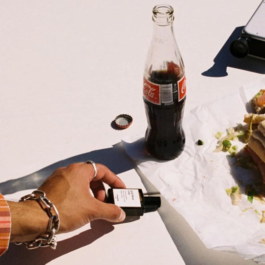Versatile Paris God Bless Cola perfume bottle held in hand beside cola bottle and sandwich on table