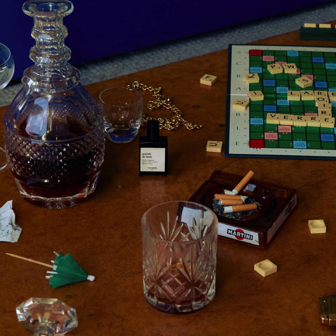 Versatile Paris Gueule de Bois perfume bottle on table with decanter, whiskey glasses, ashtray, and board game setup