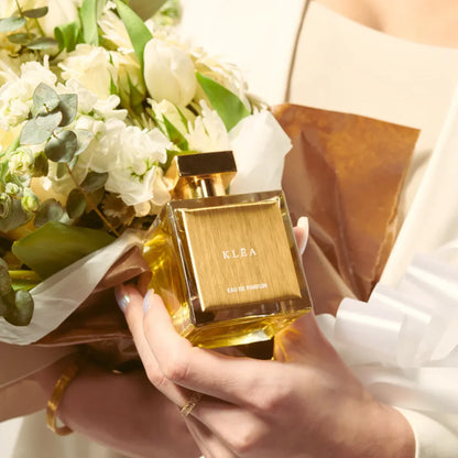 KLEA Eau de Parfum by House of Brandt luxury perfume bottle with gold cap held beside elegant white floral bouquet