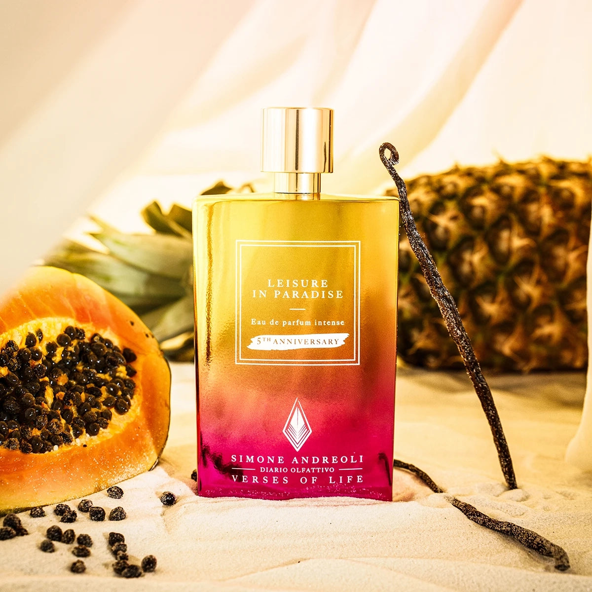 Simone Andreoli Leisure in Paradise 5th Anniversary Eau de Parfum bottle surrounded by tropical fruits, papaya, pineapple and vanilla pods on sandy background
