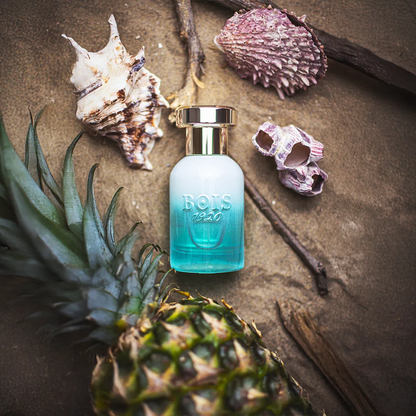 Bois 1920 Marasma perfume bottle with frosted blue gradient glass placed among pineapple, seashells and driftwood, evoking a tropical marine fragrance atmosphere.