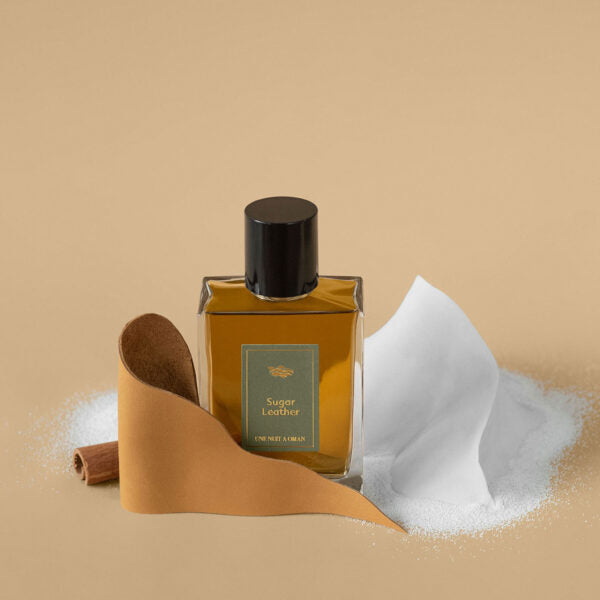 Une Nuit Nomade Sugar Leather niche perfume with leather strip, sugar crystals, and cinnamon beside the amber fragrance bottle, highlighting a warm sweet leather scent with gourmand spicy notes
