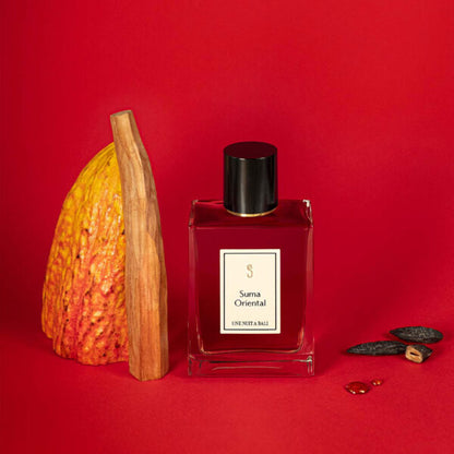 Une Nuit Nomade Suma Oriental niche perfume with cacao pod, aromatic wood, and dark resin beside the amber fragrance bottle, evoking a rich oriental scent with warm spicy and gourmand notes