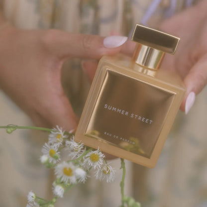 Summer Street Eau de Parfum by House of Brandt held in hand with delicate white flowers, luxury amber bottle with gold cap lifestyle fragrance image